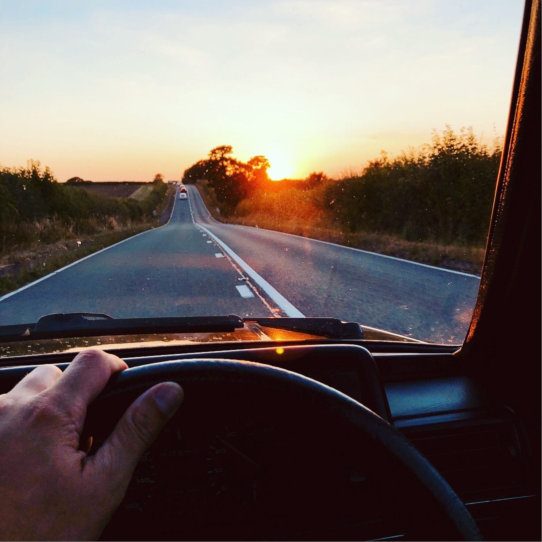 Sunset on an open B-road, seen from the driver's seat with a hand on the wheel